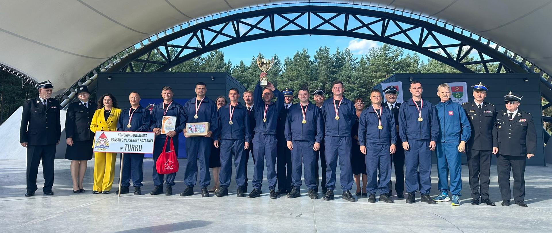 Zawodnicy wraz z osobami wręczającymi nagrody pozują do zdjęcia stojąc w amfiteatrze. Zawodnicy trzymają w rękach otrzymane nagrody oraz puchar. 