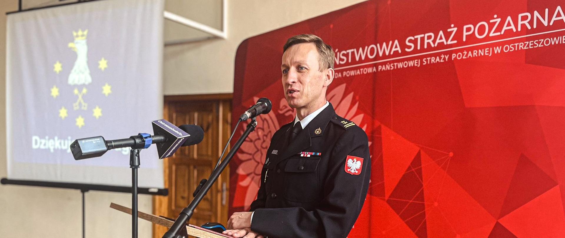 strażak stoi przed mównicą, przemawia, za nim czerwone sło z napisem państwowa straż pożarne