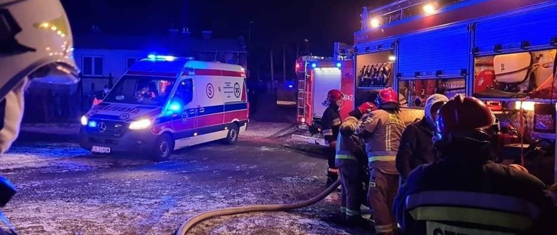 Na zdjęciu widać strażaków chwilę po akcji gaszenia pożaru rozbierających się z aparatów ochrony układu oddechowego, stojących przy samochodzie pożarniczym. W tle widać karetkę pogotowia ratunkowego na sygnałach świetlnych