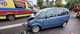 Zdjęcie przedstawia samochód osobowy marki Opel z uszkodzonym prawym przodem. Samochód stoi na środku jezdni. Przez pojazdem leżą uszkodzone elementy samochodu. Za pojazdem stoi karetka pogotowia ratunkowego. 