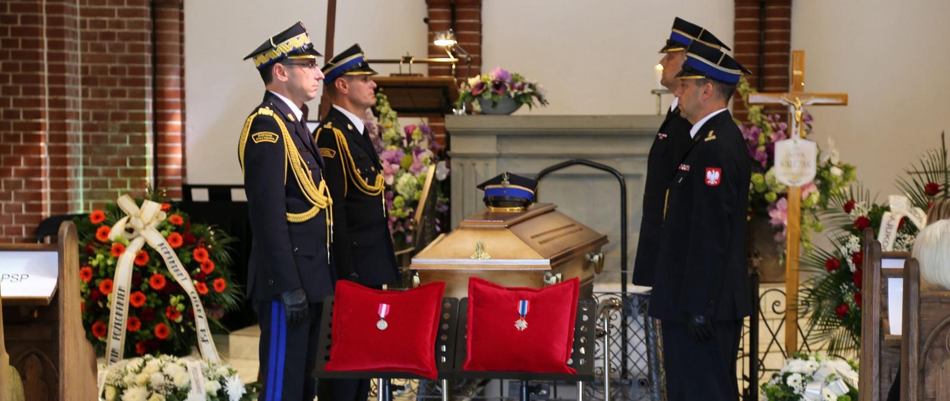 Trumna przykryta rogatywką otoczona przez strażaków pełniących wartę honorową w kościele. Przed trumną widoczne są poduszki z odznaczeniami.