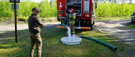 Na tle lasu stoi cięzki samochód pożarniczy. Za nim strażak, który sprawdza możliwość poboru wody ze zbiornika podziemnego. Obok zbiornika leży waż ssawny. Na pierwszym planie znajduje się leśniczy.