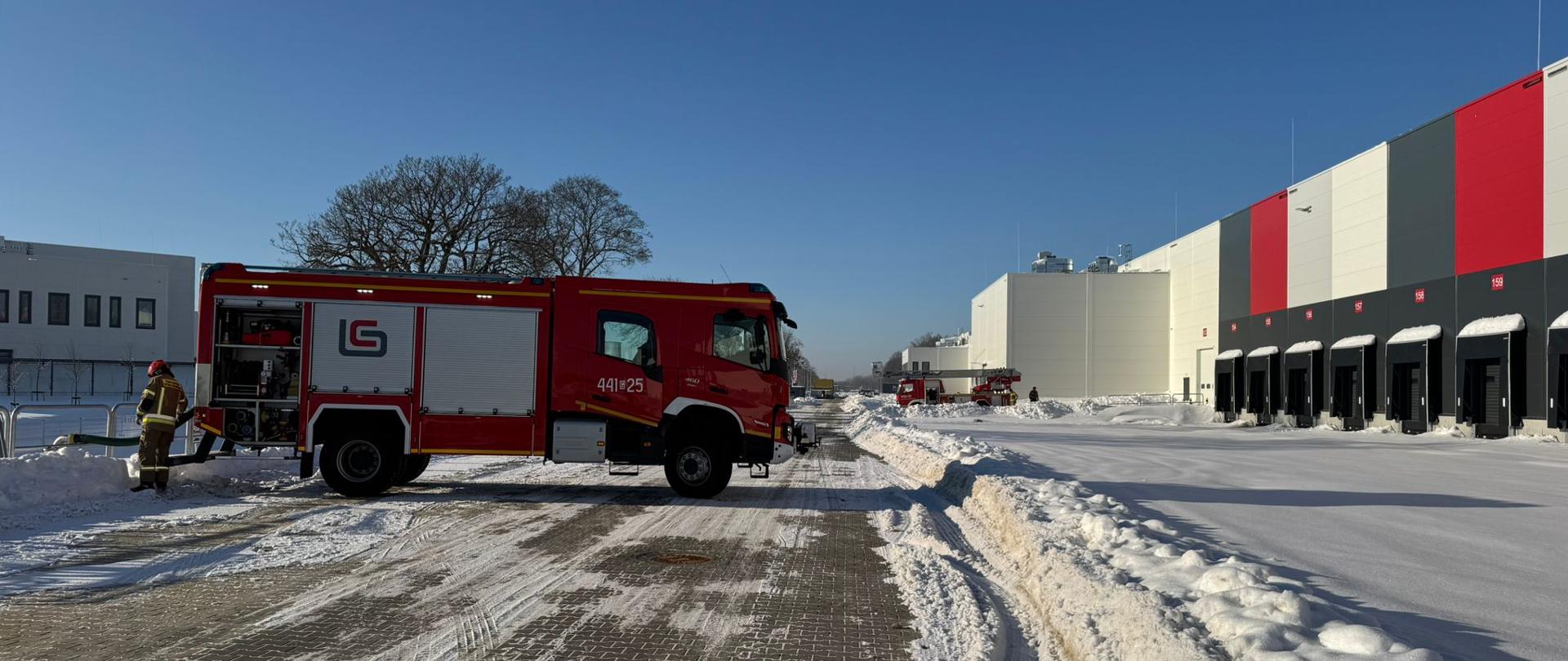 Na zdjęciu widać samochód pożarniczy, w tle drabina. Stoją na ośnieżonym placu przy obiekcie magazynowym.