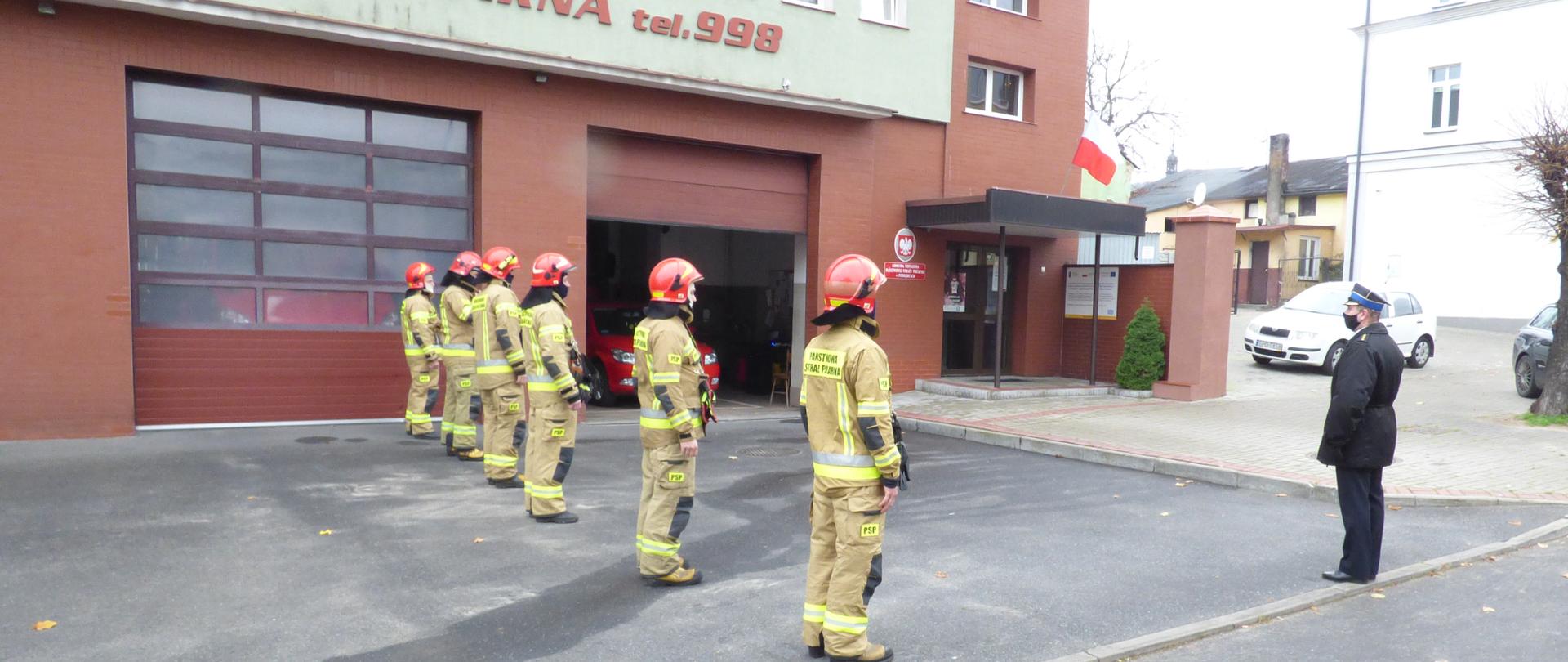 Przed budynkiem Komendy Powiatowej Państwowej Straży Pożarnej w Poddębicach. Po lewej stronie stoi sześciu funkcjonariuszy na baczność. Ubrani w strój bojowy koloru khaki oraz czerwone hełmy. Odwróceni są tyłem do zdjęcia. Po prawej stronie przodem do budynku komendy stoi komendant powiatowy Radosław Cioeślikowski ubrany w czarną kurtkę wyjściową oraz mundur. NA twarzy ma maseczkę . Brama od barażu jest otwarta, w tle widać czerwony samochód osobowy. Nad wejście do komendy jest daszek na którym powiewa biało czerwona flaga.