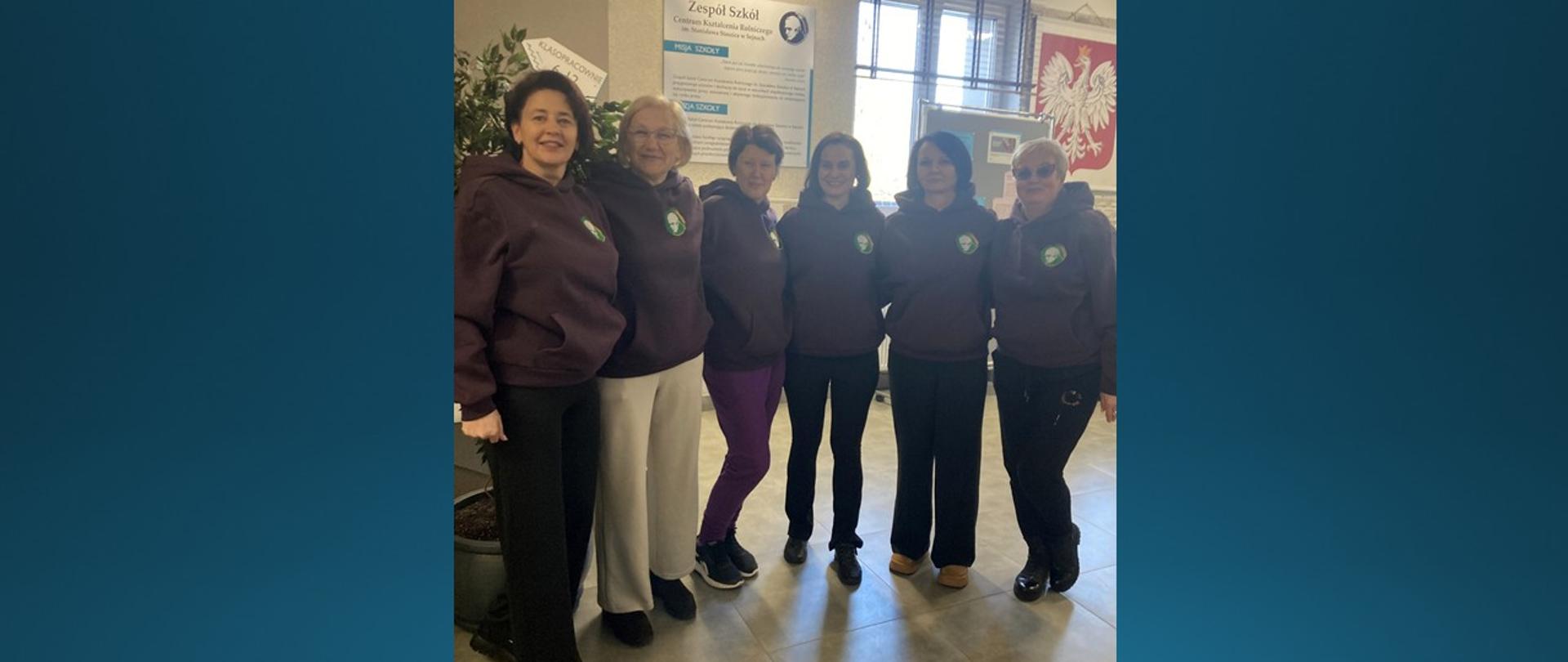 Sześć uśmiechniętych kobiet, stojących w rzędzie, ubranych w brązowe bluzy sportowe z logo szkoły.