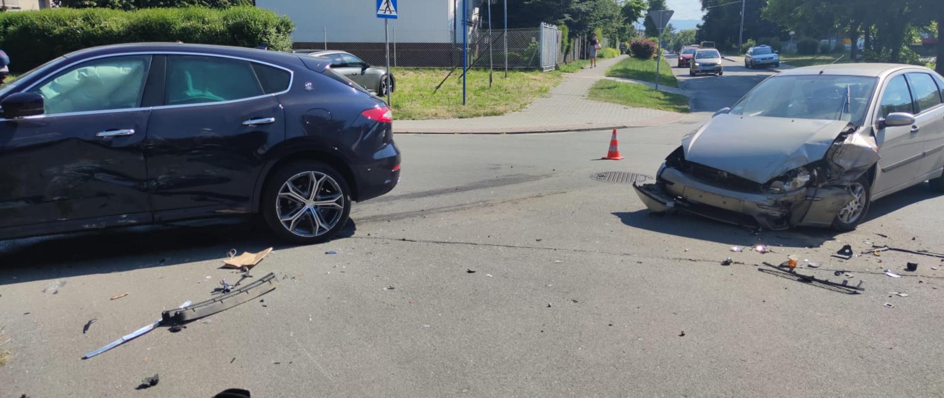 Dwa rozbite samochodu osobowe jeden ciemnego koloru a drugi jasnego stoją na skrzyżowaniu w poprzek.