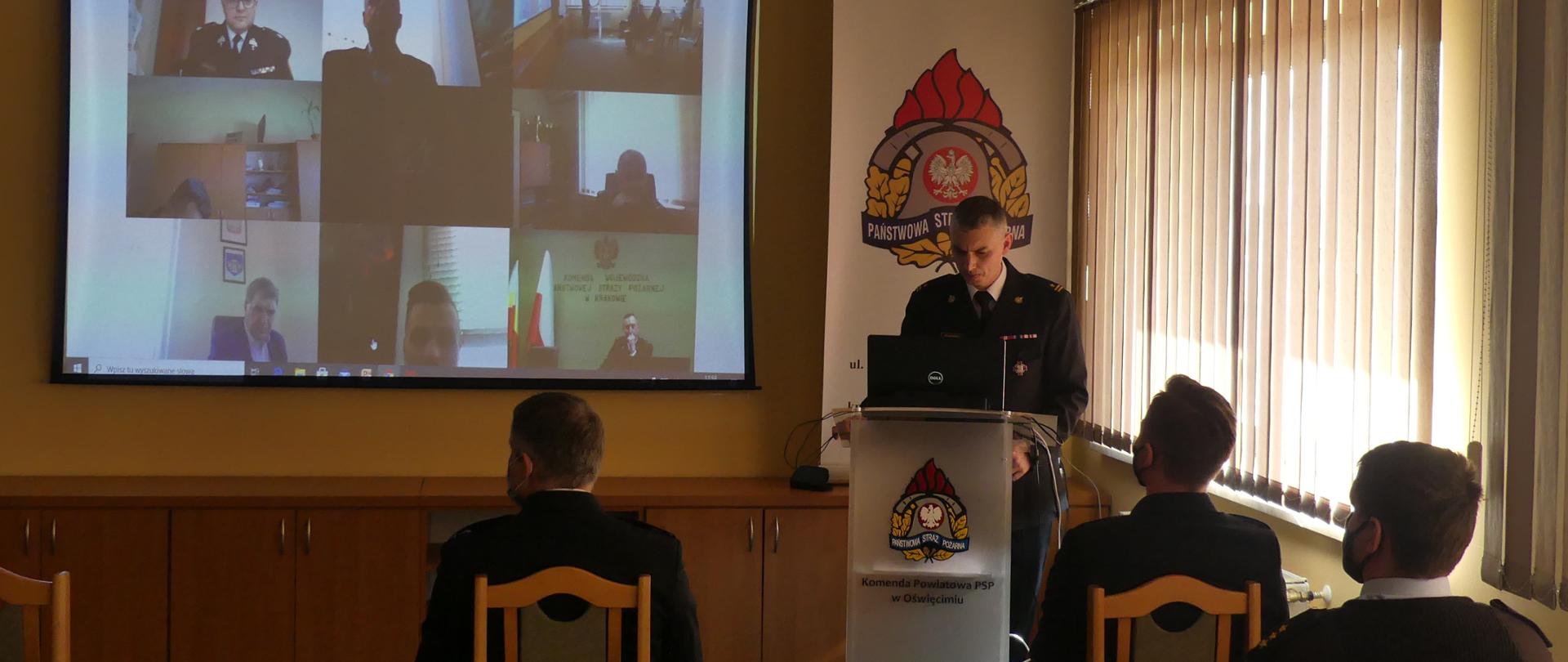 Komendant Powiatowy PSP w Oświęcimiu przy mównicy. Trzech funkcjonariuszy słuchający przemówienia. W tle ekran projektora na którym widać sylwetki uczestników wideokonferencji.