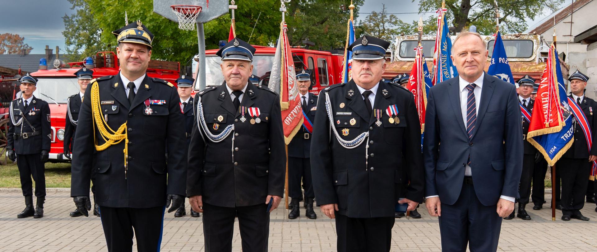 Czterej mężczyźni — trzej w mundurach strażackich oraz jeden w eleganckim garniturze — stoją w jednym rzędzie na wybrukowanym placu. Za ich plecami znajdują się strażacy trzymający sztandary oraz pojazdy pożarnicze.