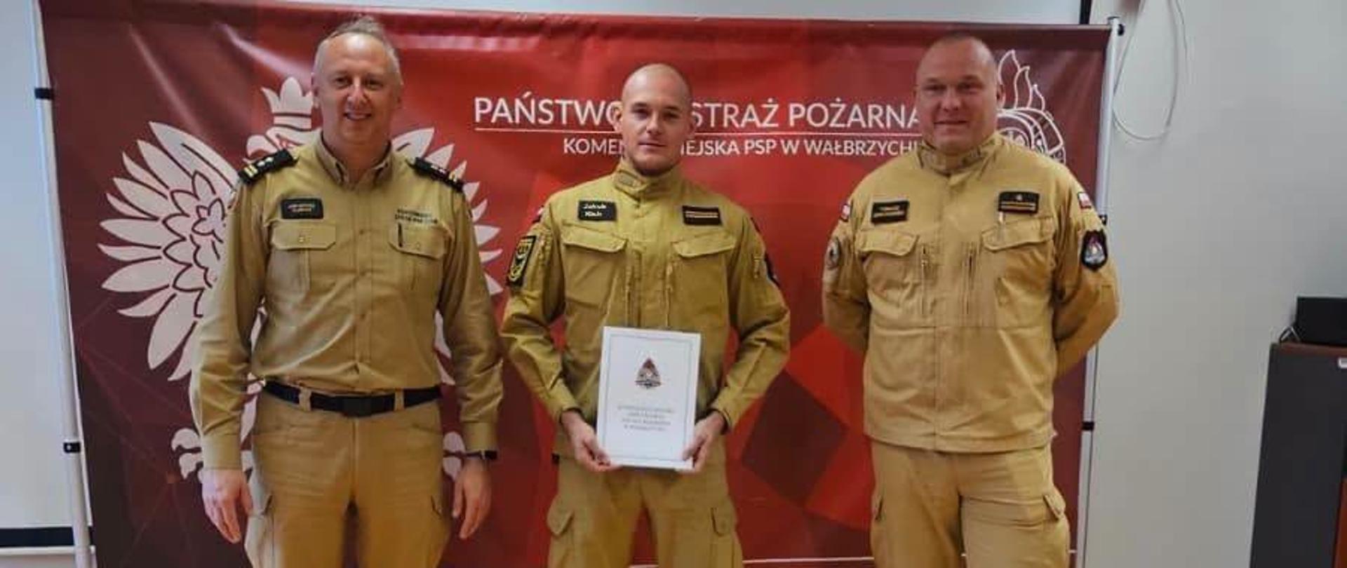 Trzech mężczyzn w piaskowych mundurach służbowych Państwowej Straży Pożarnej stoi w rzędzie w jasnym pomieszczeniu. Za nimi znajduje się czerwony baner z godłem Polski oraz napisem "Państwowa Straż Pożarna, Komenda Miejska PSP w Wałbrzychu". Mężczyzna w środku trzyma oburącz oprawiony dyplom, natomiast dwaj pozostali stoją po jego bokach z rękami splecionymi za plecami.
