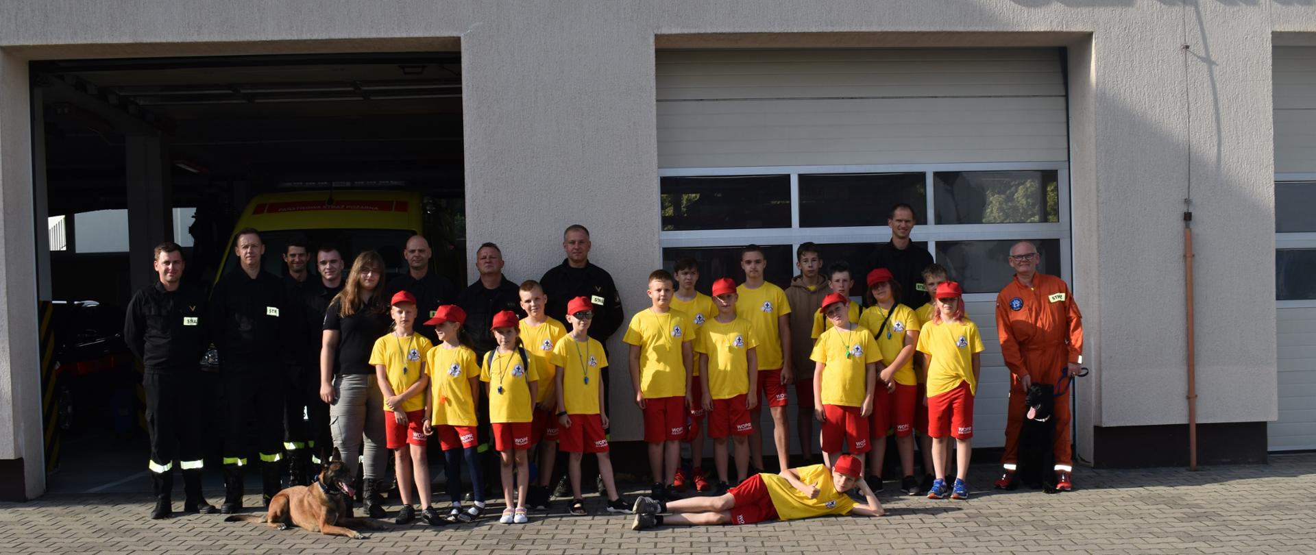 Na zdjęciu grupa dzieci oraz strażacy i przewodnicy z psami ratowniczymi na tle pleszewskiej komendy straży.s