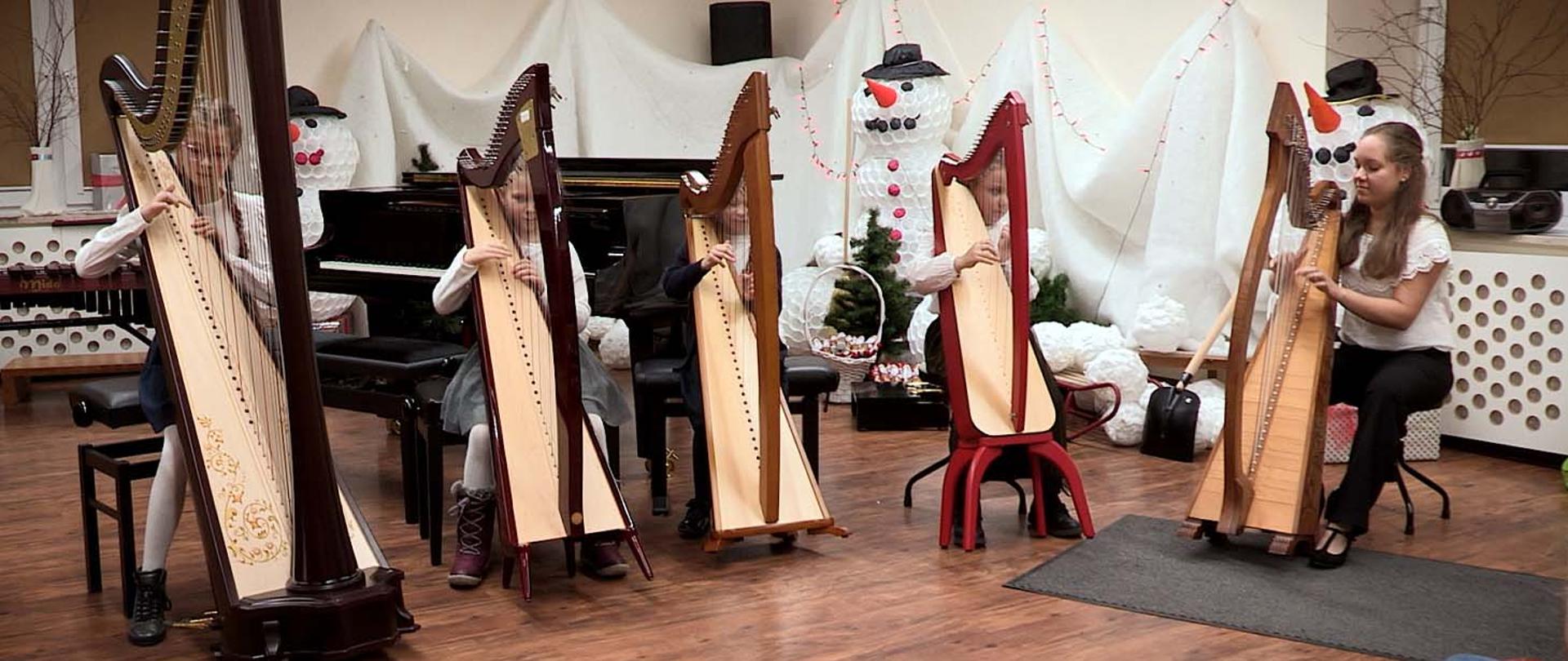 Na zdjęciu cztery dziewczynki i jedna dorosła kobieta grają na harfach. Znajdują się w pomieszczeniu, za nimi stoi czarny fortepian a za nim, przy ścianie białe dekoracje świąteczne przypominające bałwany.