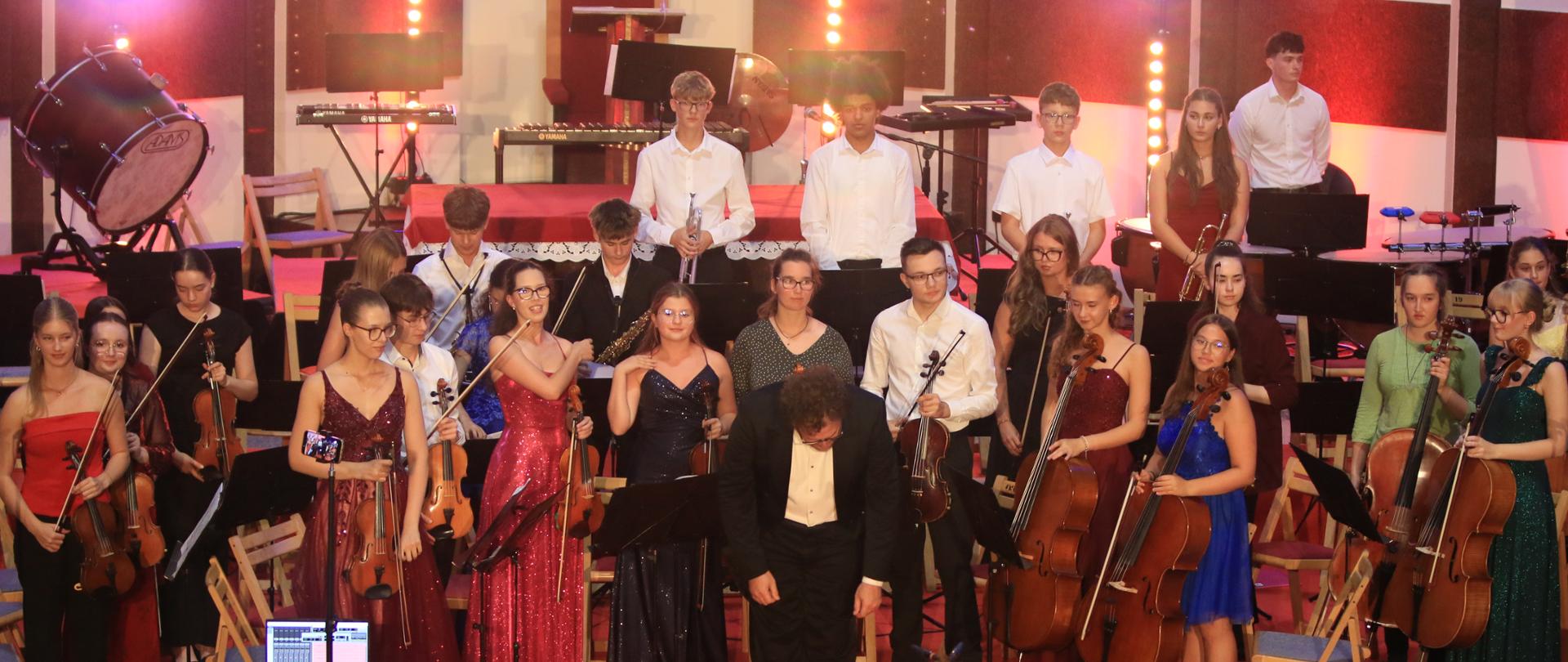 Młodzieżowa orkiestra symfoniczna, ubrana w eleganckie stroje wieczorowe. Na pierwszym planie widać sekcję smyczkową ze skrzypcami i wiolonczelami, a w tylnych rzędach instrumenty perkusyjne i dęte. Na środku sceny dyrygent w czarnym fraku kłania się nisko przed siedzącą w ławkach publicznością.