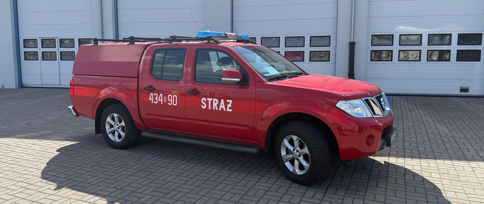 Czerwony Nissan Navara z numerami operacyjnymi. Samochód strażacki stojący przed jednostką i garażami.