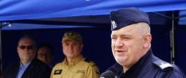 Funkcjonariusz Policji stoi przed mównicą i przemawia, za nim stoją zaproszeni goście.