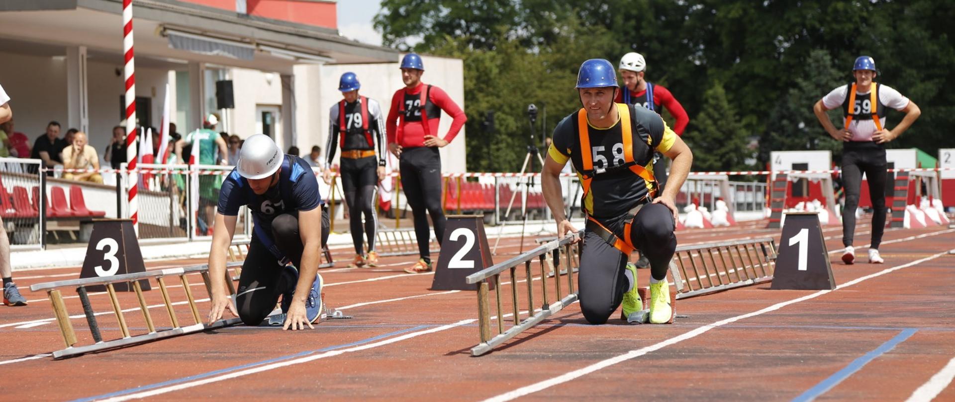Dwaj zawodnicy przygotowani do startu w konkurencji drabiny hakowej. W tle widoczni inni zawodnicy przyglądający się zmaganiom 