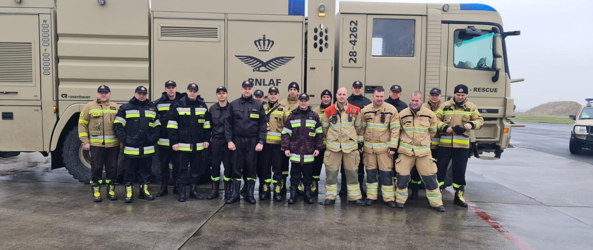 Strażacy stoją na tle pojazdu piaskowego pojazdu gaśniczego należącego do holenderskiej wojskowej straży pożarnej.