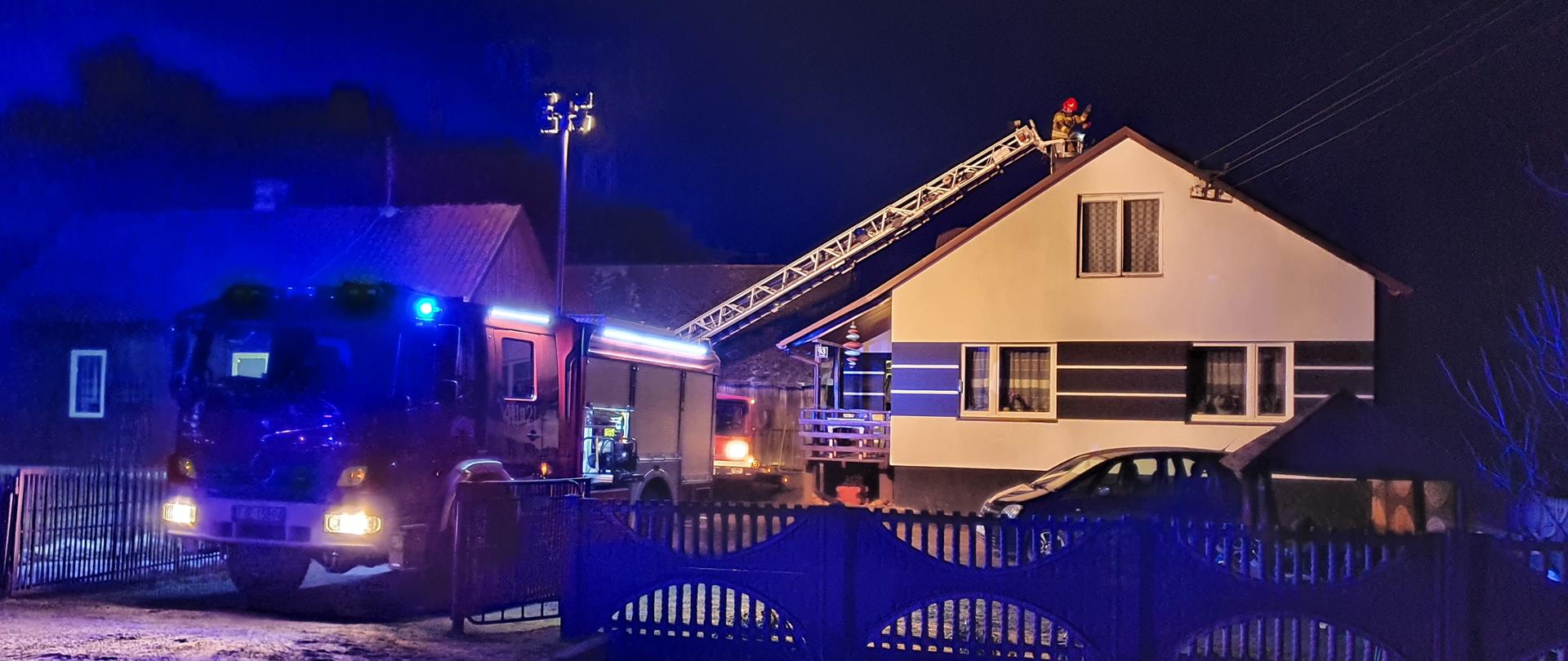 Zdjęcie przedstawia budynek mieszkalny, parterowy. Po lewej stronie znajduje się samochód ratowniczo-gaśniczy z włączoną alarmową sygnalizacją świetlną oraz włączonymi światłami mijania. Na dachu samochodu rozłożony jest masz oświetleniowy. W tle widnieje rozłożony park drabinowy. W jego koszu znajduje się strażak w umundurowaniu specjalnym z czerwonym hełmem na głowie. Strażak pracuje przy obtrącaniu sadzy w przewodzie kominowym.