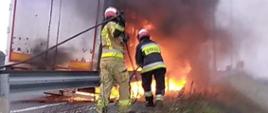 Naczepa objęta pożarem, dwóch strażaków przygotowujących się do podania prądu gaśniczego.