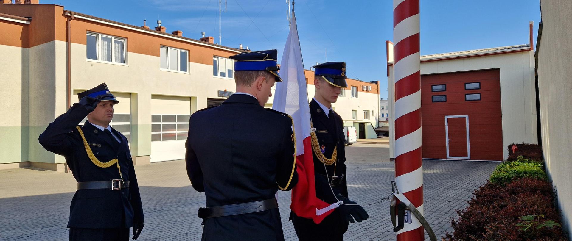 Uroczyste podniesienie flagi państwowej, na zdjęciu poczet flagowy przygotowuje się do podniesienia, w tle budynki Komendy