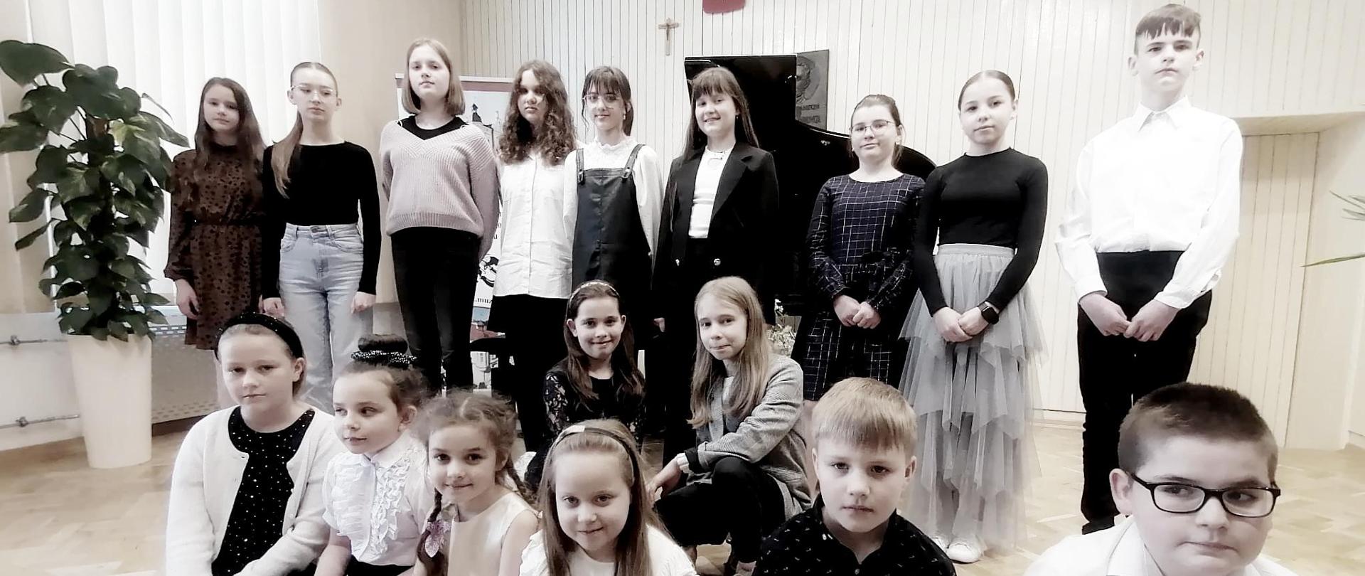 Zdjęcie uczestników Szkolnego Konkursu Interpretacji Muzycznej przedstawiające chłopców i dziewczynki w strojach galowych ustawione na scenie auli.