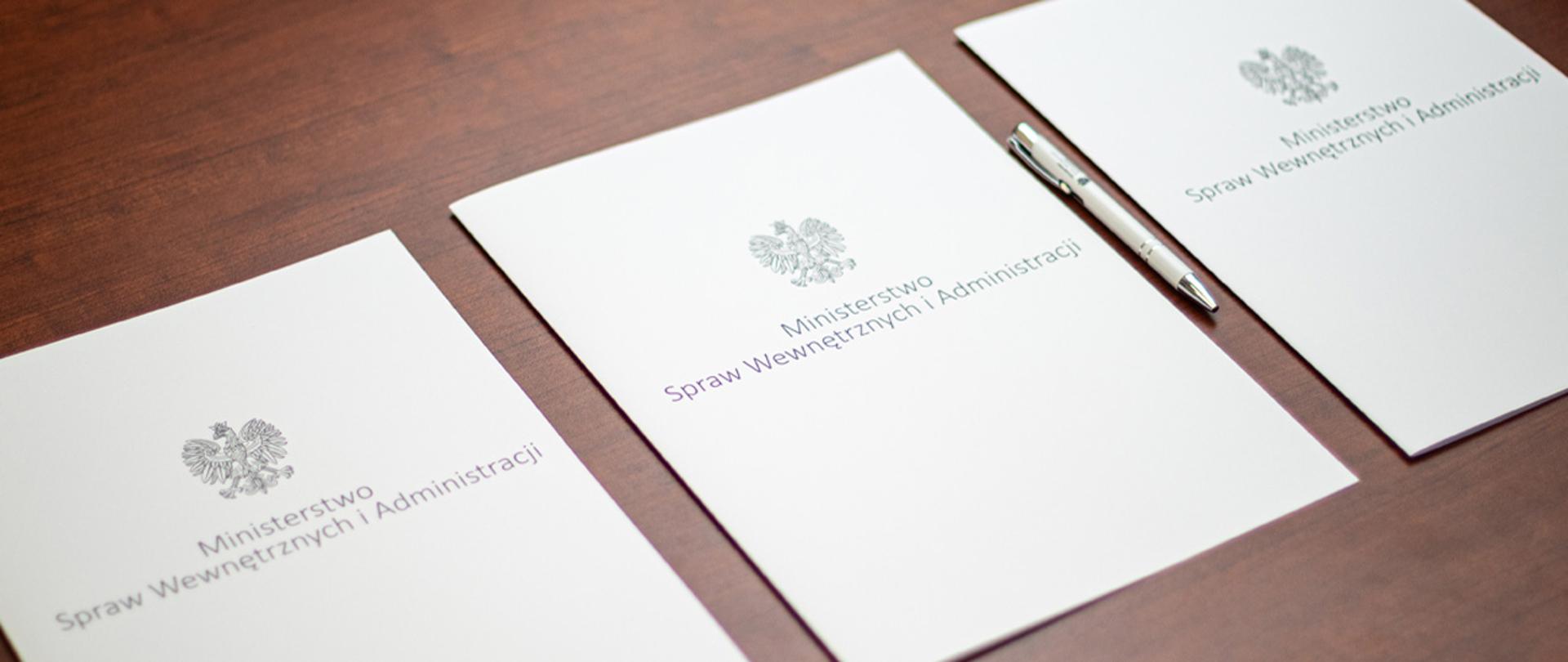 Na zdjęciu widać trzy leżące w rzędzie teczki na dokumenty z napisem "Ministerstwo Spraw Wewnętrznych i Administracji" i orłem z godła narodowego. Obok jednej z nich leży długopis MSWiA.