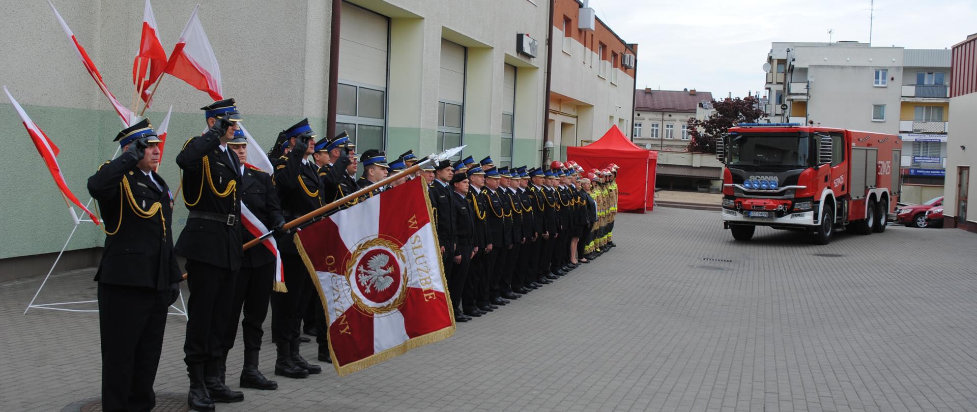 Zdjęcie przedstawia strażaków Komendy podczas uroczystości. W galowych mundurach, Ustawieni w szeregu, na pierwszym planie sztandar Komendy, w tle samochód pożarniczy, flagi państwowe.