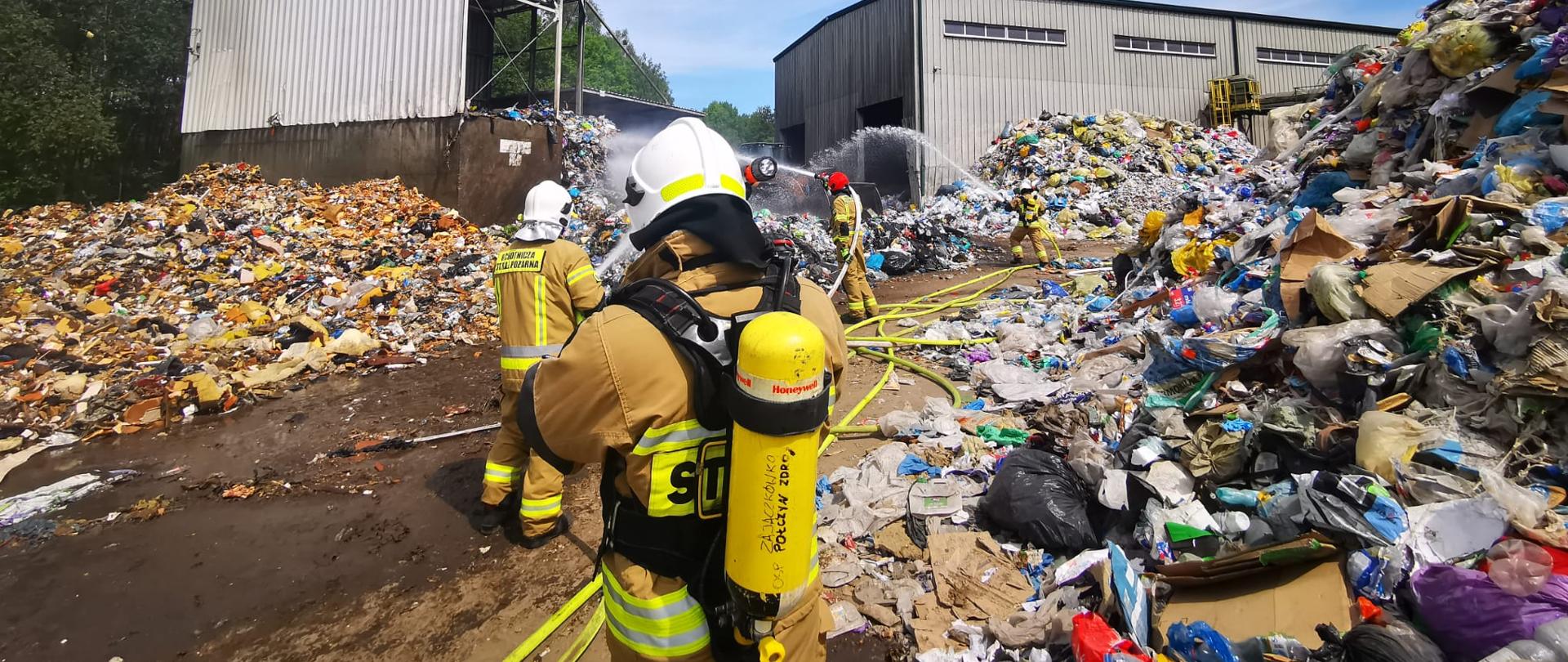 Zdjęcie przedstawia gaszenie składowiska odpadów na wysypisku śmieci przez strażaków.