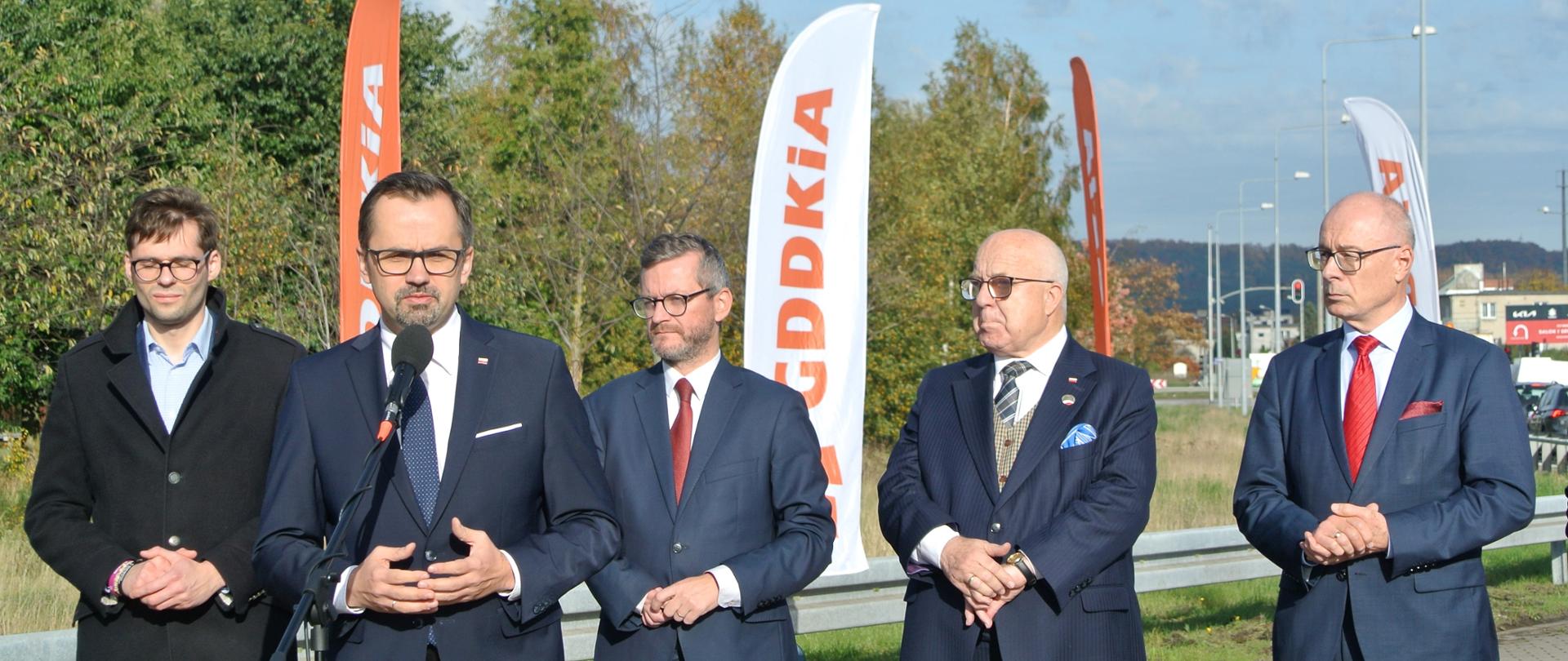 Pięciu mężczyzn stoi obok siebie w jednym rzędzie. Drugi z lewej stoi wiceminister Marcin Horała i mówi do mikrofonu.