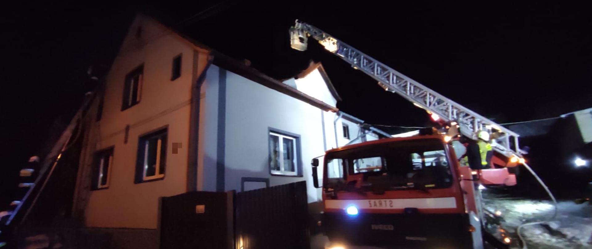 Zdjęcie wykonane w porze nocnej. Zdjęcie przedstawia drabinę mechaniczną sprawioną przy budynku mieszkalnym (w bramie gospodarstwa). W tle po lewej stronie widoczna część budynku. Skrajnie po lewej stronie widoczna drabina przystawna oparta o szczyt budynku, na której znajduje się strażak w ubraniu specjalnym i białym hełmie na głowie.