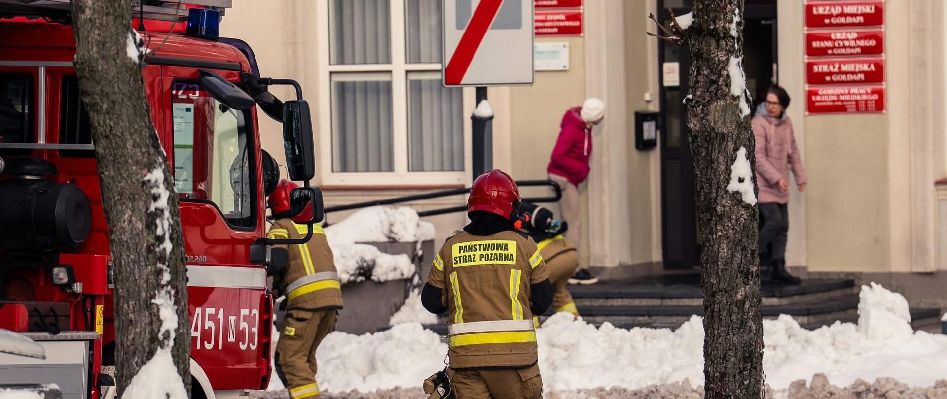 Zdjęcie przedstawia część pojazdu pożarniczego oraz trzech funkcjonariuszy Komendy Powiatowej Państwowej Straży Pożarnej w Gołdapi zlokalizowanych przed wejściem do obiektu Urzędu Miejskiego w Gołdapi podczas sprawdzenia organizacji oraz warunków ewakuacji z budynku