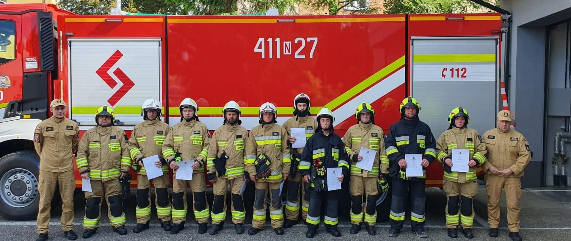 Na tle dużego samochodu strażackiego stoi grupa strażaków w mundurach i hełmach.