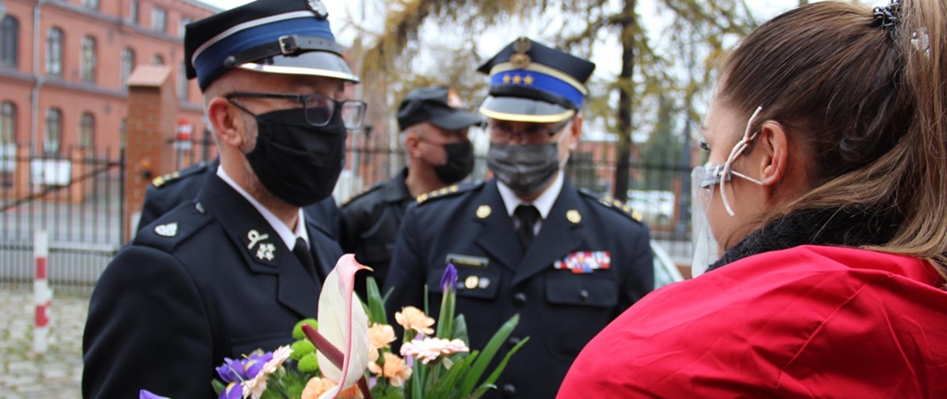 Na zdjęciu Prezes OSP Raciążek wręcza kwiaty ii dziękuje przedstawicielce Pani minister w resorcie Rolnictwa i Rozwoju Wsi. On w umundurowaniu wyjściowym. Wydarzenie ma miejsce na placu, przed siedzibą KW PSP w Toruniu. W tle kujawsko-pomorski komendant wojewódzki PSP.