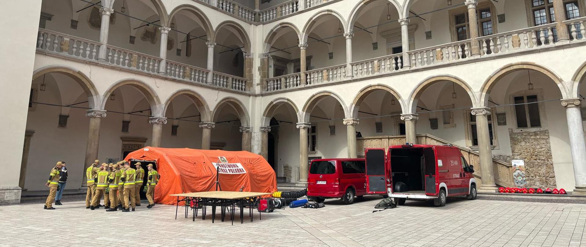 Dziedziniec Zamku Królewskiego na Wawelu. Na wykonanym z jasnego kamienia podłożu rozstawiony pomarańczowy namiot pneumatyczny, przy którym stoi grupa strażaków w umundurowaniu koloru piaskowego, po prawej stronie dwa czerwone samochody (bus i dostawczy), na środku placu dużych rozmiarów stół z jasnym blatem (płytą OSB)