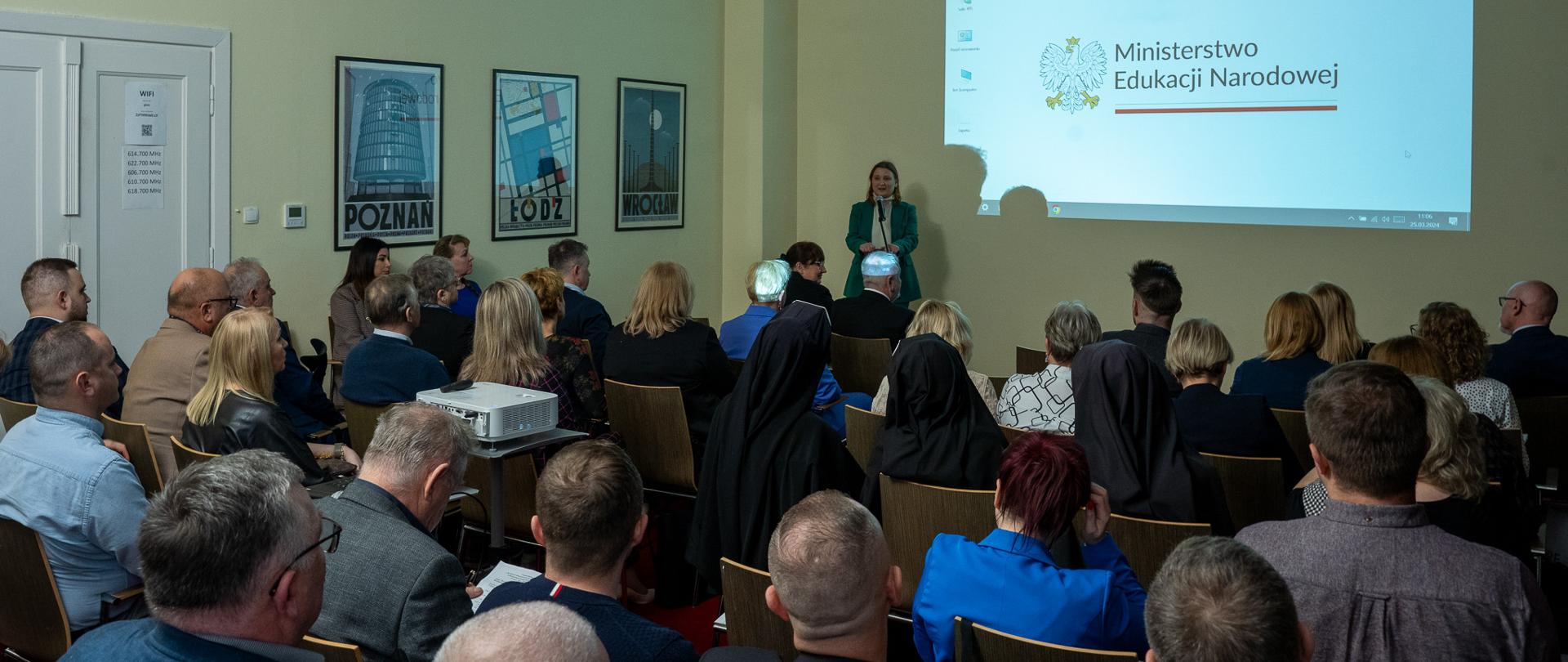 Minister Paulina Piechna-Więckiewicz wygłasza przemówienie. Przed nią ustawiony jest mikrofon na statywie. Uczestnicy spotkania siedzą na wprost na krzesłach i przysłuchują się wypowiedzi minister. Za nią na ścianie projektor wyświetlił logo Ministerstwa Edukacji Narodowej. Po lewej stronie na ścianie wywieszone są trzy plakaty z wizerunkiem miasta Poznań, Łódź i Wrocław. 