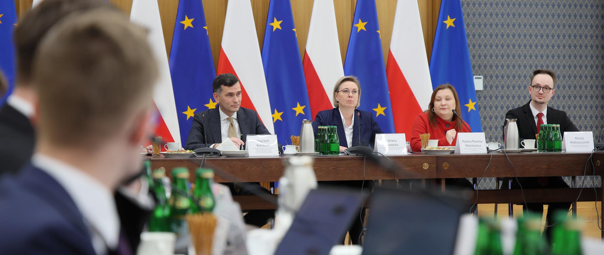 Zdjęcie sytuacyjne. Na zdjęciu uczestnicy posiedzenia Rady Dialogu z Młodym Pokoleniem obradują przy stole konferencyjnym. Na dalszym planie, widoczni są od lewej: Wojciech Kuraszyk, Zastępca Dyrektora w Departamencie Społeczeństwa Obywatelskiego w KPRM; Adriana Porowska, Sekretarz Stanu w KPRM i Wiceprzewodnicząca Komitetu do spraw Pożytku Publicznego, Paulina Piechna-Więckiewicz, Podsekretarz Stanu w Ministerstwie Edukacji Narodowej oraz Maciej Waleszczyński, Sekretarz Rady do spraw Polityki Młodzieżowej, Ministerstwo Edukacji Narodowej. W tle widoczne są flagi Polski i Unii Europejskiej