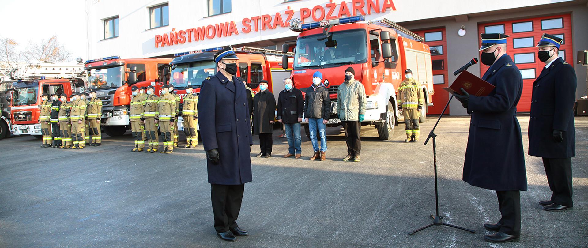 Uroczystości odejścia na emeryturę funkcjonariusza Komendy odbywające się na tle budynku komendy, wozów strażackich oraz strażaków