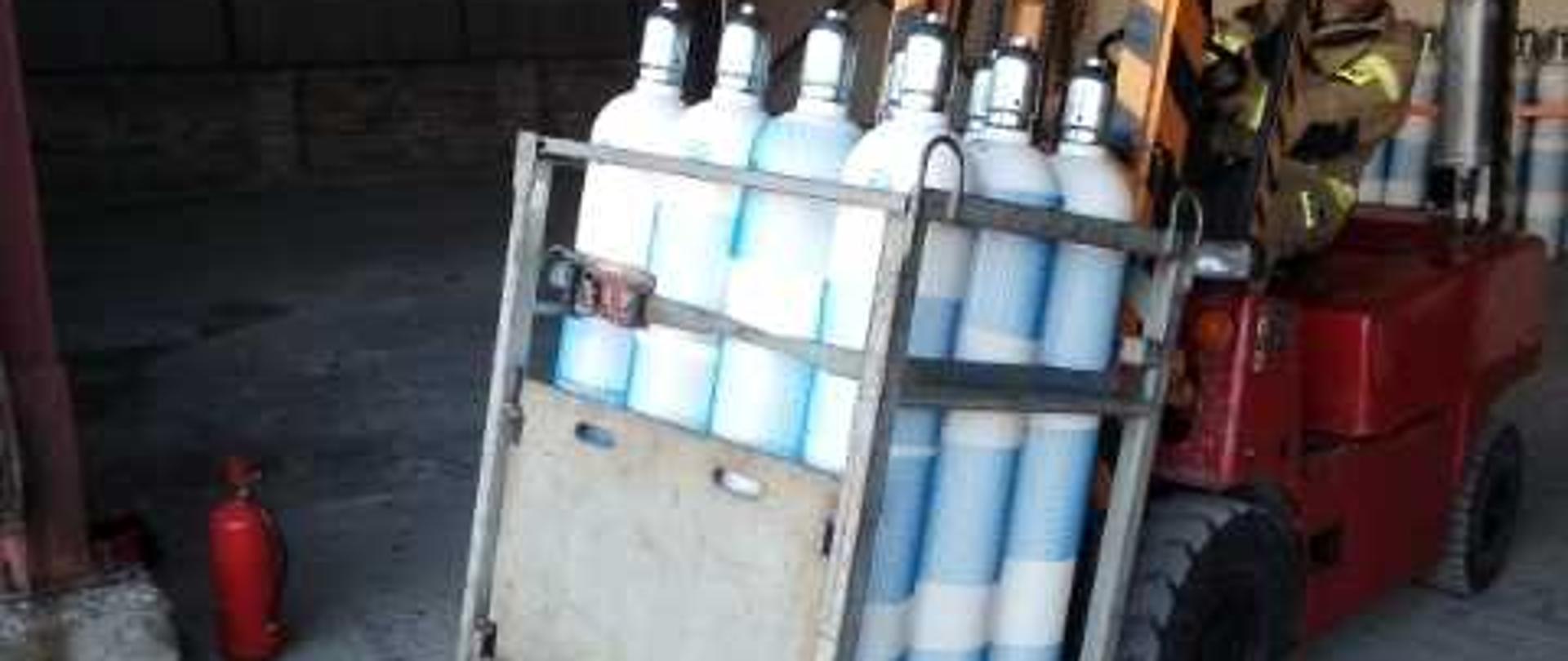 Zdjęcie przedstawia wózek widłowy prowadzony przez strażaka. Na wózku znajduje się specjalny kosz transportowy w którym umieszczone są butle zawierające tlen medyczny. Strażak ubrany w ubranie specjalne w kolorze żółtym. Wózek widłowy jest w kolorze czerwonym. 