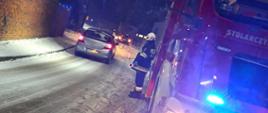 Na drodze stoi wóz straży pożarnej (widoczny jest jego przód i prawy bok), a obok niego jeden ze strażaków w ubraniu specjalnym w kolorze ciemnym i w białym hełmie. W oddali widać innego strażaka i samochód osobowy, który uderzył w drzewo. Na drodze leży śnieg i stoją pachołki ostrzegawcze. Pora nocna. Obok wozu strażackiego przejeżdża srebry pojazd osobowy.
