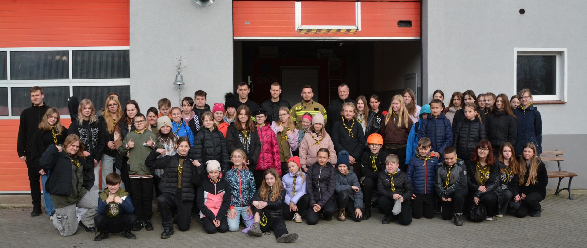 Zdjęcie wykonano w ciągu dnia na placu Komendy Powiatowej Państwowej Straży Pożarnej w Sępólnie Krajeńskim. Na zdjęciu grupa kilkudziesięcioro dzieci oraz młodzieży z drużyny harcerskiej pozuje razem ze strażakami do wspólnego zdjęcia. W tle budynek garaży.