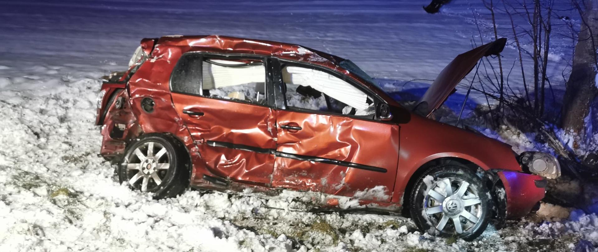 Zdjęcie przedstawia pomarańczowy samochód osobowy po zderzeniu bocznym. Cały bok auta jest rozbity, widać wystrzelone poduszki powietrzne w tym boczne. W tle widać pole pokryte śniegiem, zdjęcie zrobione nocą.