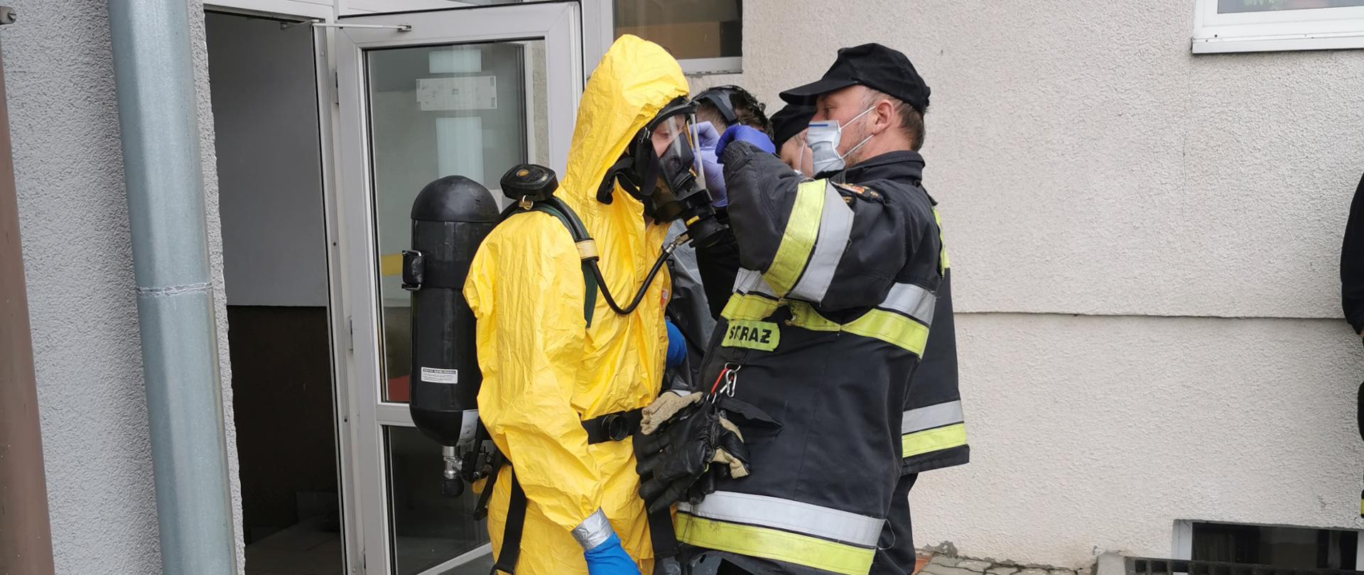 Na pierwszym planie dwóch strażaków, strażak z lewej w ubraniu ochronnym chemicznym koloru żółtego, na twarz maska, na plecach aparat ochrony układu oddechowego. Strażak z prawej w kurtce ubrania specjalnego koloru granatowego, na twarzy maseczka chirurgiczna. Strażak z prawej sprawdza dopięcia kombinezonu chemicznego. W tle siwa elewacja budynku JRG w Szubinie. 