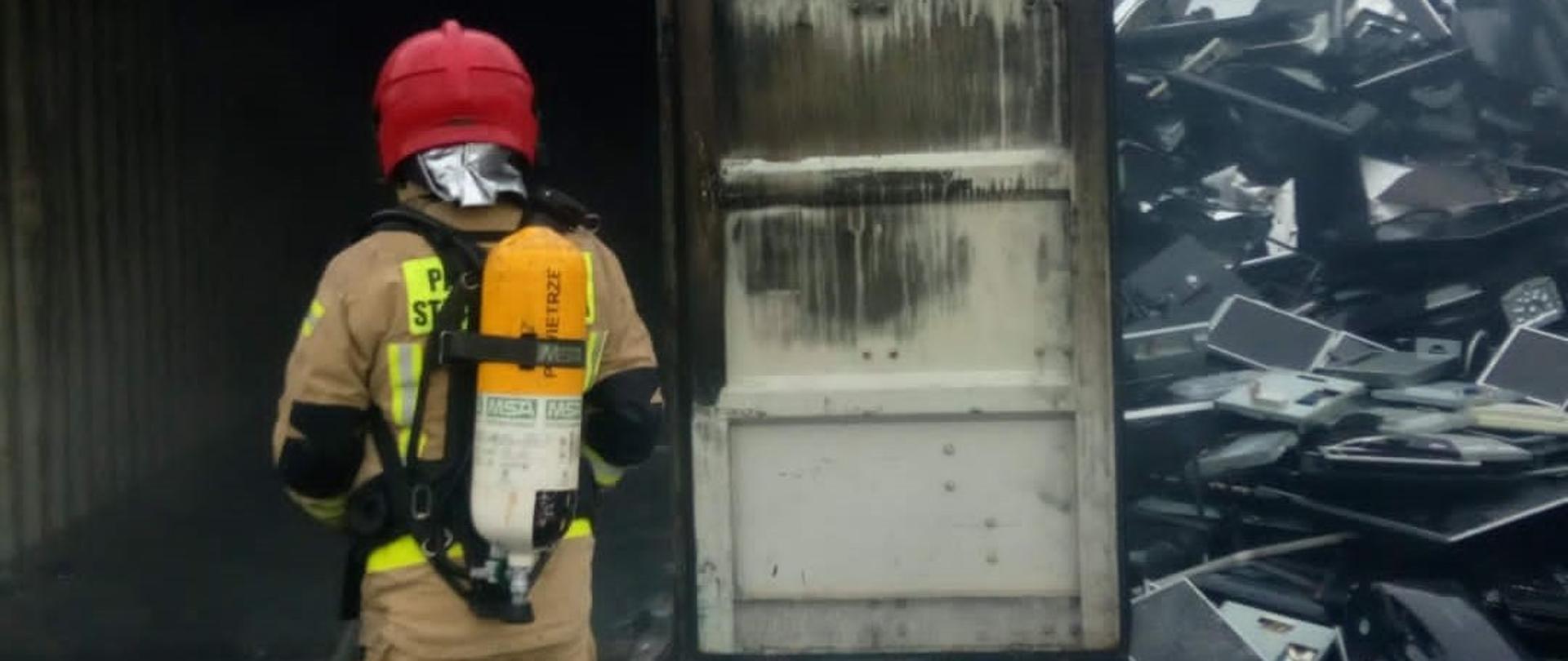 Zdjęcie przedstawia strażaka zabezpieczonego w aparat ochrony dróg oddechowych, który z linią gaśniczą wchodzi do metalowego kontenera. Po prawej stronie widać pryzmę odbiorników telewizyjnych przeznaczonych do recyklingu. 