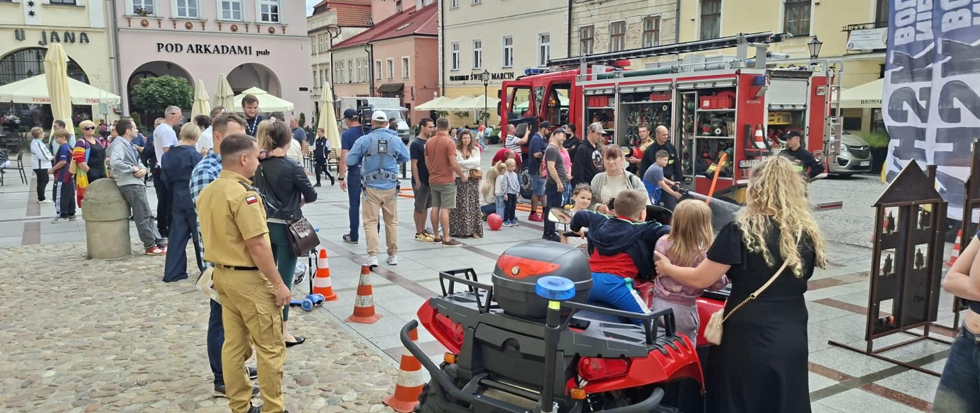 Grupa mieszkańców Tarnowa, w tym dzieci, ogląda sprzęt strażacki podczas akcji edukacyjnej na rynku. Widoczny wóz gaśniczy, quad ratowniczy i funkcjonariusze PSP prowadzący pokazy.