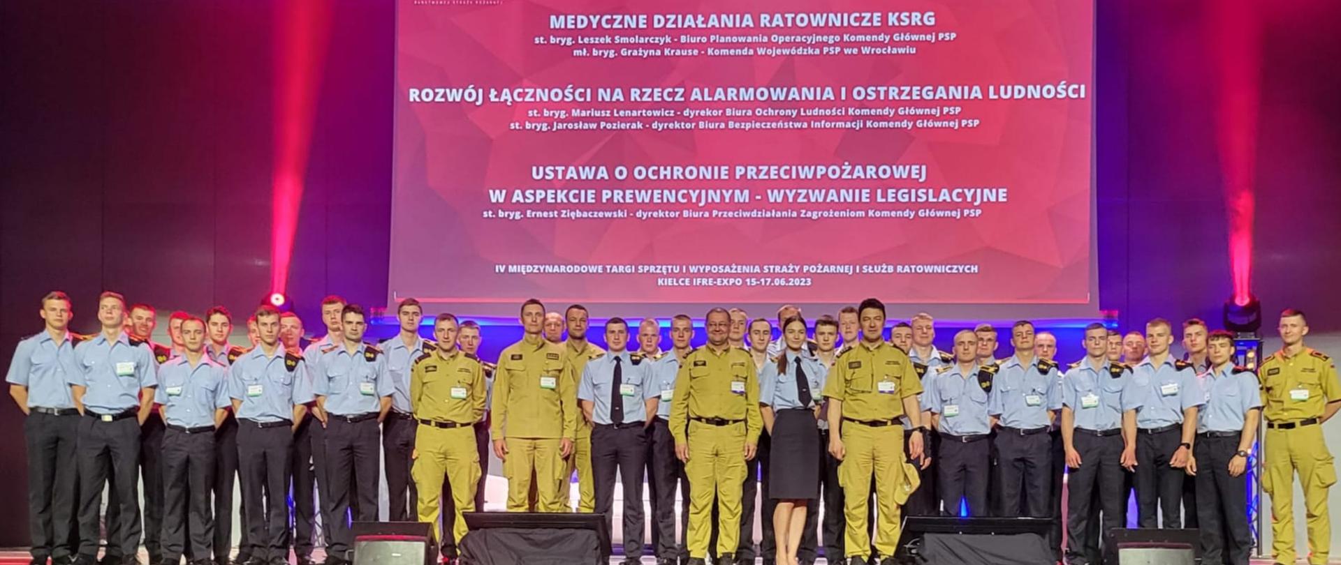 Grupowe zdjęcie kierownictwa Centralnej Szkoły PSP wraz z kadetami i kadrą Szkoły stojących na scenie za nimi wyświetlony slajd koloru czerwonego z białymi napisami KONFERENCJA i nazwy bloków tematycznych „Medyczne działania ratownicze w KSRG”, „Rozwój łączności na rzecz alarmowania i ostrzegania ludności” „Ustawa o ochronie przeciwpożarowej w aspekcie prewencyjnym – wyzwanie legislacyjne”