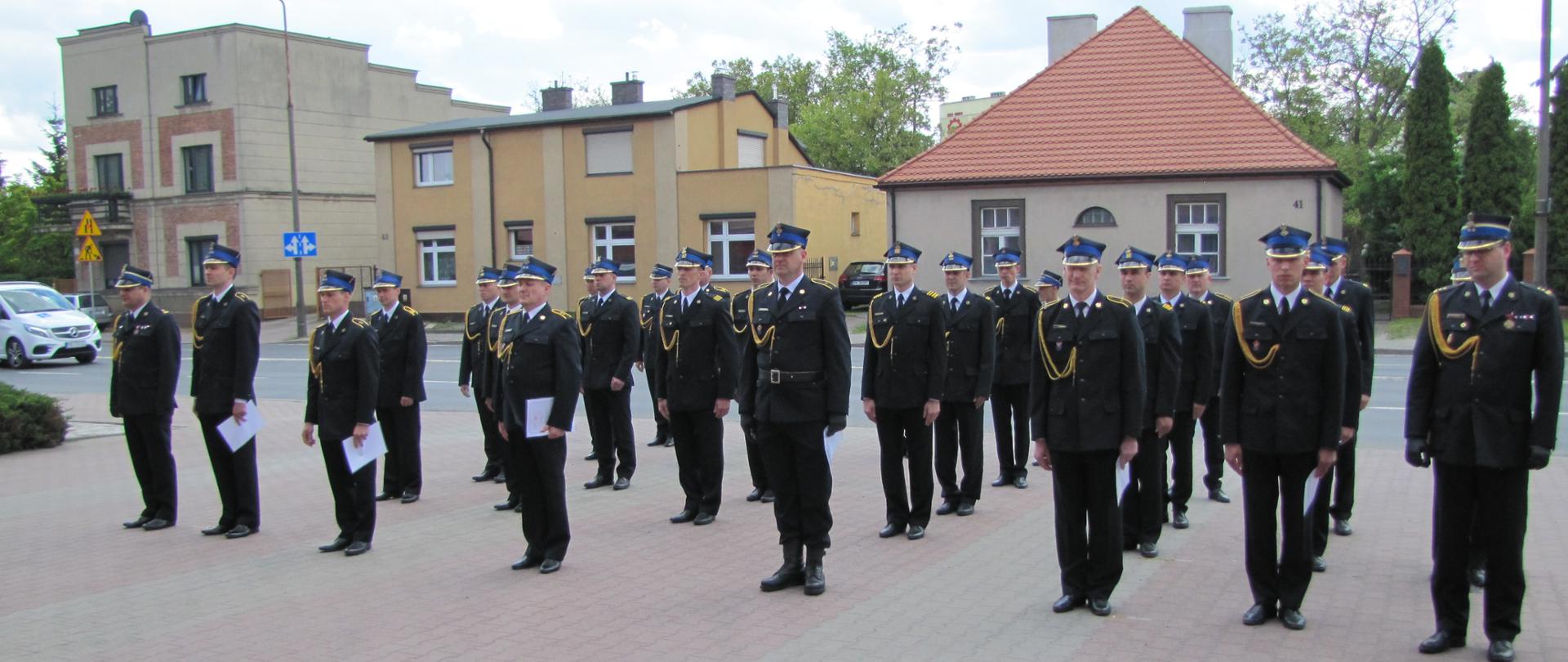 Na zdjęciu widzimy strażaków stojących przodem do kadru w kilku rzędach. Ubrani w ciemne mundury, ciemne buty, czapki na głowach, widoczne są także złote sznury na mundurach. Mężczyźni w pierwszym rzędzie trzymają białe kartki. W tle widzimy zabudowania mieszkalne, samochody przejeżdżające po ulicy. Strażacy stoją na utwardzonym placu z kostki brukowej.