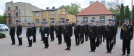Na zdjęciu widzimy strażaków stojących przodem do kadru w kilku rzędach. Ubrani w ciemne mundury, ciemne buty, czapki na głowach, widoczne są także złote sznury na mundurach. Mężczyźni w pierwszym rzędzie trzymają białe kartki. W tle widzimy zabudowania mieszkalne, samochody przejeżdżające po ulicy. Strażacy stoją na utwardzonym placu z kostki brukowej.
