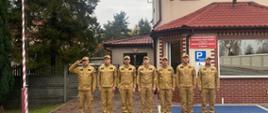 Strażacy na tle budynku KP PSP Pajęczno śpiewają hymn Polski pod masztem