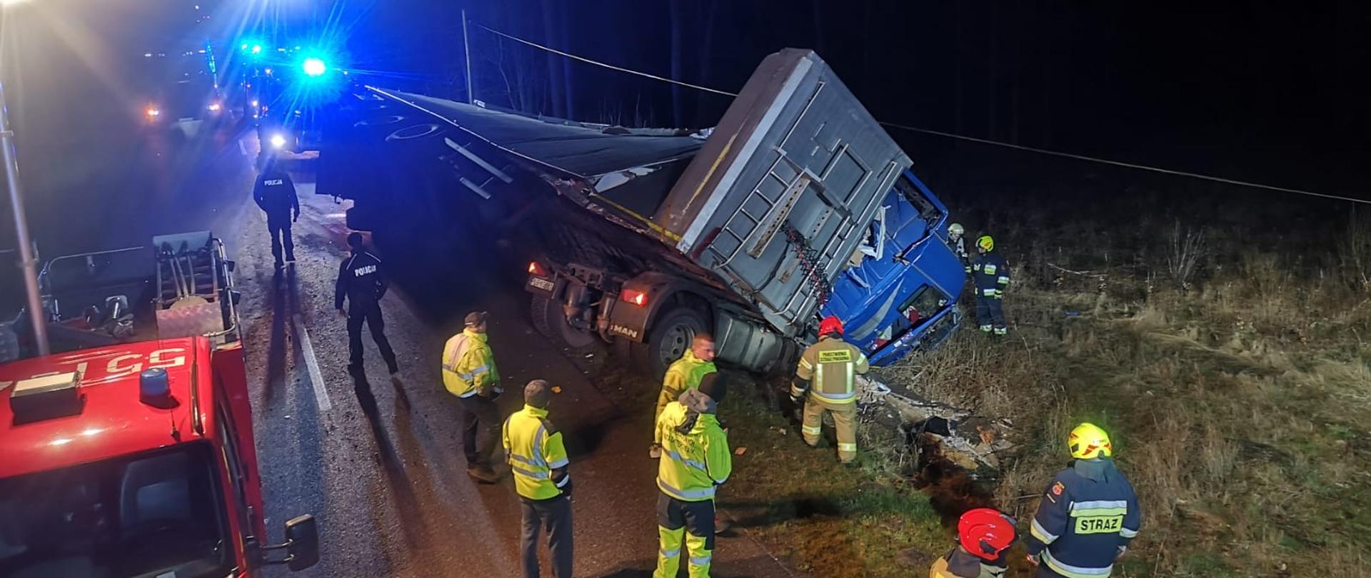 Zdjęcie przedstawia drogę na której na jednym pasie częściowo leży przewrócony samochód ciężarowy z naczepą. Teren oświetlany jest z pojazdu straży pożarnej najaśnicą. Na zdjęciu znajdują się strażacy oraz ratownicy medyczni.