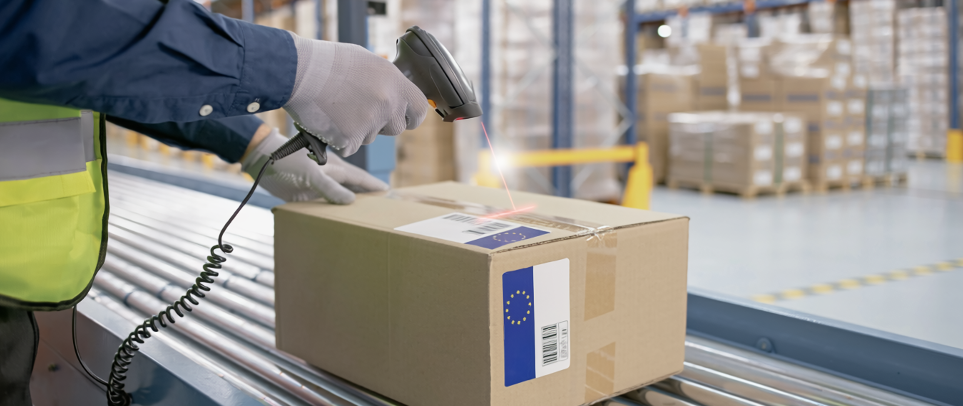 Osoba w kamizelce odblaskowej skanuje kod kreskowy na kartonie, leżącym na taśmie transportowej w magazynie pełnym regałów z zapakowanymi przesyłkami.