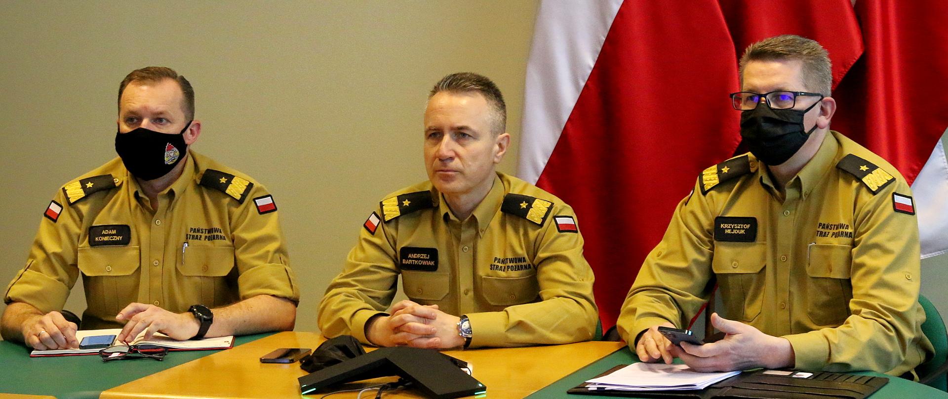 Po lewej stronie zastępca komendanta głównego PSP nadbryg. Adam Konieczny, następnie komendant główny PSP, nadbryg. Andrzej Bartkowiak, po prawej stronie zastępca KG PSP nadbryg. Krzysztof Hejduk. Wszyscy trzej siedzą przy stole podczas wideokonferencji, ubrani są w nowe umundurowanie służbowe koloru "musztardowego"